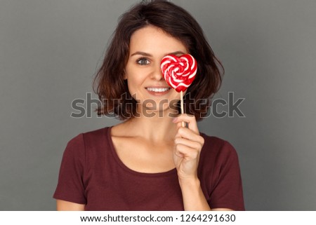 Young woman standing isolated on grey wall covering eye with heart shaped lollipop looking camera smiling cheerful close-up