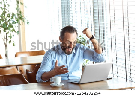 Mature man trader sitting at table at cafe at daytime with cup of coffee looking at laptop screen hand in fist up success achieved smiling happy.
