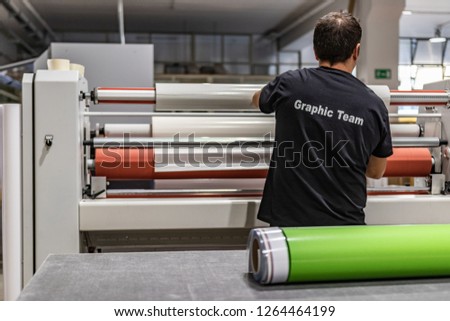Specialized staff working big vinyl rolls on a professional printing machine and laminator. Details of the preparation and paper feed. White tubes, industrial workplace, occupation concept.  Royalty-Free Stock Photo #1264464199