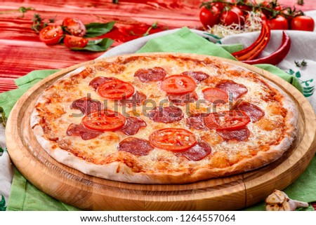 Pizza pepperoni on a round wooden board on a red wooden background, decorated with napkins, chili pepper and cherry tomatoes. close-up. top view. flat lay