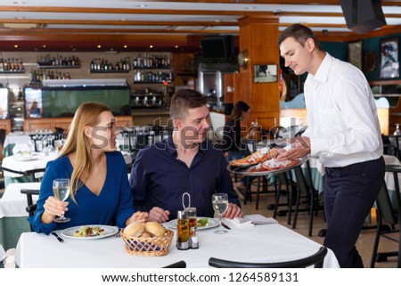 Cheerful waiter serving dishes to adult couple having dinner at restaurant