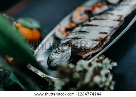 Grill fried trout fish with fried shrimps on the metal steel plate with mandarin, green leafs, magnolia flowers and leafs and orange persimmon nearby on the black background. Shallow focus on fish.