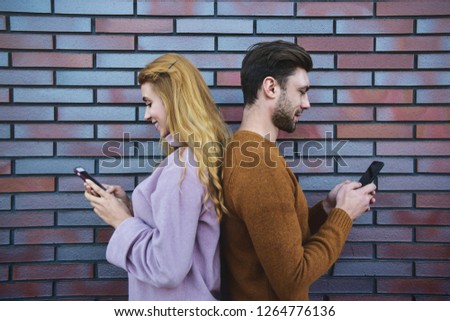 Side portrait of beautiful young couple is using smartphones and smiling, standing back to back against brown brick wall.