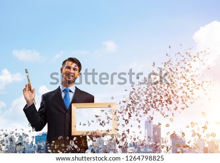 Conceptual image of young and successful businessman in black suit holding paintbrush in hand and smiling while standing against modern cityscape view with flying letters on background.
