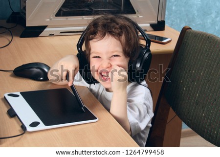 Little 2 3 year old baby girl in white clothers draws at the home computer in graphics drawing tablet. The child is holding a pen and laughs. On the head are huge headphones. close up.