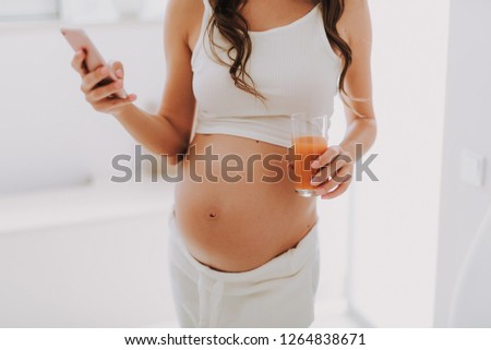 Cropped photo of young woman holding cellular and multivitamin juice in hand while standing in light room