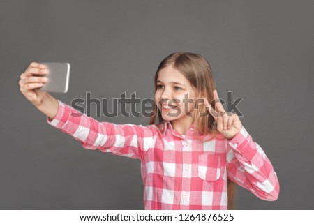 Little girl standing isolated on grey wall taking selfie photo on smartphone showing peace gesture smiling happy to camera