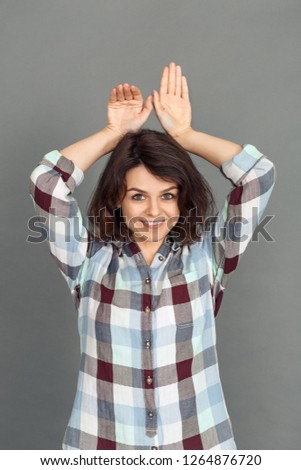 Young woman making bunny ears with hands standing isolated on grey wall looking camera smiling playful