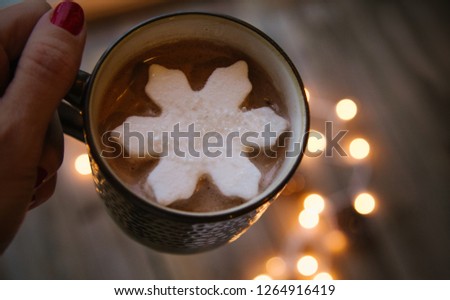 A woman's hand holding a mug of hot cocoa with a snowflake marshmallow and lights in the background.