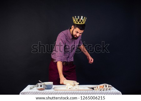 a handsome baker man with beard confectioner bakery with a golden crown on his head king of the kitchen bakes bread , pizza, pasta holding a rolling pin kneading dough in studio on a black background