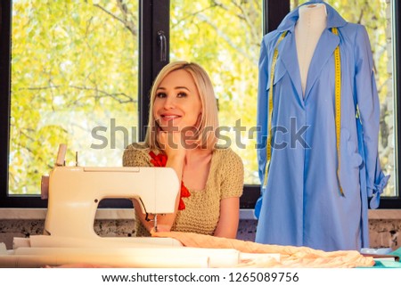 beautiful blonde female seamstress writes curve template. tailor creates a collection outfits sews clothes sewing machine in workshop. young woman designer clothes notes ideas sitting near window