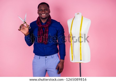 African American man tailor seamstress workshop stylish male model clothes designer measuring tape on neck posing next to the mannequin holding scissors in hand on pink background in the studio