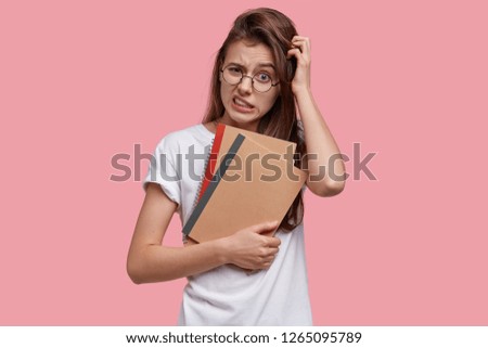 Displeased young woman scratches head, clenches teeth, looks in bewilderment, thinks about creative ideas for writing essay, holds notepads, dressed in casual clothes, isolated on pink background