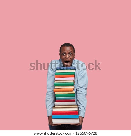 Vertical shot of stupefied black man leans head on pile of books, wears round glasses, surprised with many tasks for seminar, cramms material, models over pink studio wall with free space above