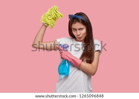 Gloomy young housewife points at muscle with serious offensive expression, angry with husband who doesnt help her with house cleaning, holds detergent, washes mirror in bathroom, poses indoor
