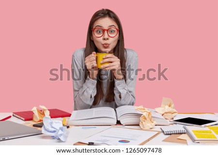 Photo of attractive young female holds mug with hot beverage, wears big spectacles, dressed in stylish shirt, poses at desktop with opened encyclopedia, isolated over pink wall. Working atmosphere