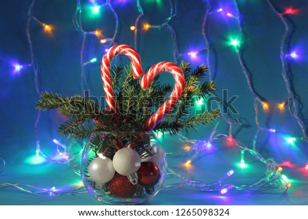 Two Christmas candies in the form of a heart, striped canes, in a vase with fir branches against the background of colored garlands