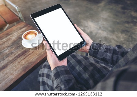 Mockup image of woman's hands holding black tablet pc with blank screen with coffee cup on wooden table in cafe