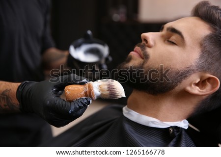 Professional hairdresser applying shaving foam onto client's skin in barbershop Royalty-Free Stock Photo #1265166778
