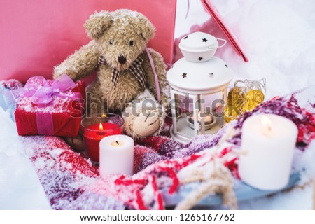 A close-up of a teddy bear with Christmas candles on a cozy checkered plaid outdoors next to a red gift box sprinkled with snow. Christmas festive mood