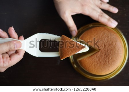 Female hand using pie cutter server to cut up Japanese cotton cheesecake in triangle shape before serving. Top view of cake on dark wooden table background. (close up, selective focus, space for text)