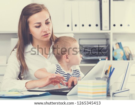Female with kid is working behind laptop in office.