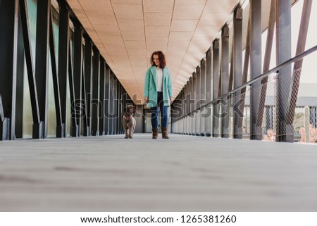 beautiful young woman smiling and standing in the middle of an aisle, using mobile phone. Indoors. taking a picture of her cute brown dog.