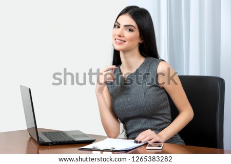 Young business woman sitting at her office and smiling at the camera while holding a pen close to her mouth.