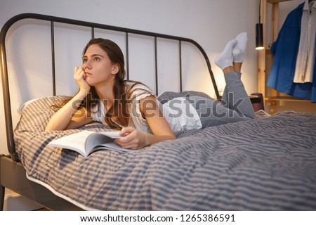 Bedtime, leisure and relaxation concept. Picture of beautiful young brunette woman in socks and night suit lying comfortably on bed with feet raised, having thoughtful dreamy look while reading book