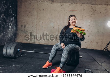 plump girl is looking at the hamburger while resting aftre workout. full length photo. girl lifting the ban