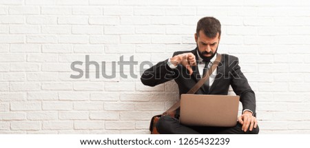 Businessman with his laptop sitting on the floor showing thumb down sign with negative expression