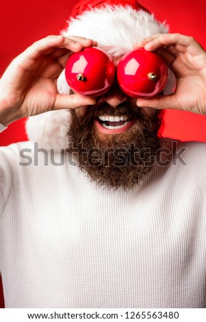 Man in Santa hat covered eyes red by Christmas ball. Christmas toys. Happy Santa hold christmas ball decoration. Happy holidays. Merry Christmas and happy New Year. Man in Santa's hat.
