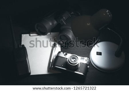 Blank page with copy space, pen, portable radio, binoculars and photo camera on a black spy agent table background. Top secret documents. Detective agent dossier. Interrogation report.