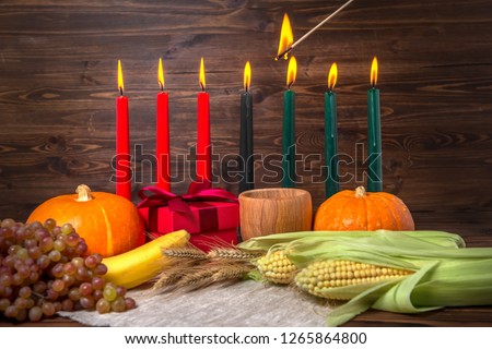 ignition of Kwanzaa traditional candles, holiday concept with gift box, pumpkins, ears of wheat, grapes, corns, banana, bowl and fruits on wooden background, close up