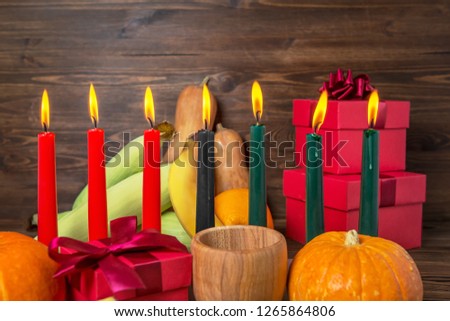 Kwanzaa festive concept with burning candles, gift box, pumpkins, ears of wheat, grapes, corns, banana, bowl and fruits on wooden background, close up