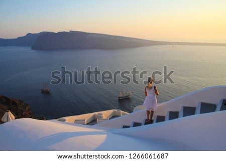 Young woman watching colorful beautiful sunset view of Mediterranean Sea, islands, boat and sea on white outdoor terrace. Female in white sundress on summer Europe travel vacation in Santorini, Greece