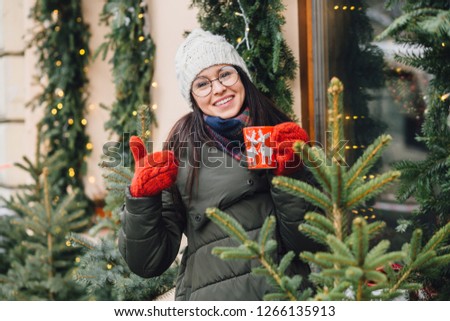 Seasonal concept. Outdoors portrait of pretty brunette business woman show thumb up drinking tea from red cup on the holiday city. Smiling, drinking coffee. Wearing coat, hat, red knitted mittens.