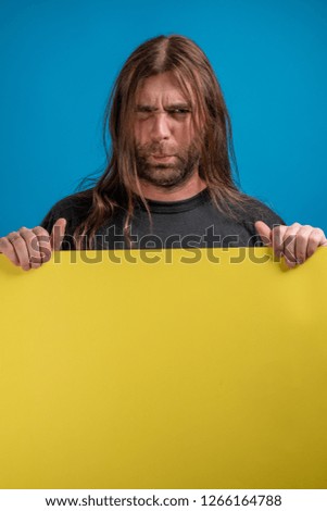 Angry male portrait making faces while holding a yellow cardboard banner. Studio shot against blue background