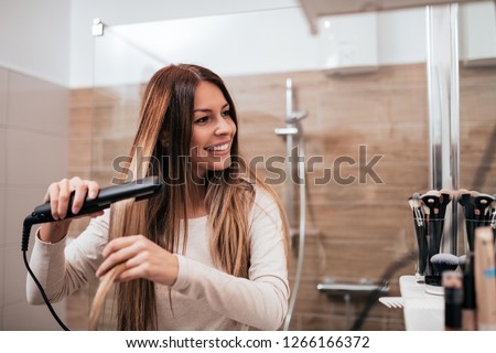 Beautiful young smiling woman using a hair straightener while looking into the mirror in bathroom. Royalty-Free Stock Photo #1266166372