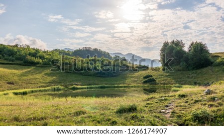 View of a mountain lake with smooth water and reflection, surrounded by grass and bushes, illuminated by the sun through the clouds. Caucasus, Russia