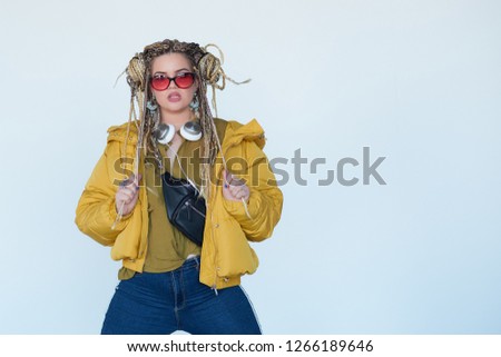 young blonde girl in sunglasses with african pigtails listens to music in headphones and dances against a white background.
