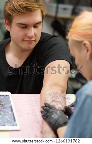 Male client of tattoo salon looking down at hand and waiting. Tattoo master drawing picture on man's skin using special instrument. Professional tattooist working in black gloves.
