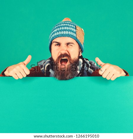 Man in warm hat shows thumbs up on green background, copy space. October and November sale idea. Autumn and cold weather concept. Hipster with beard and flirty face wears warm clothes
