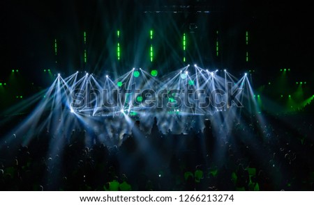 Scene illuminated by beautiful rays of lighting equipment. The concert crowd having fun at the center in the big hall. Television is broadcast live. Many people look towards the stage. 