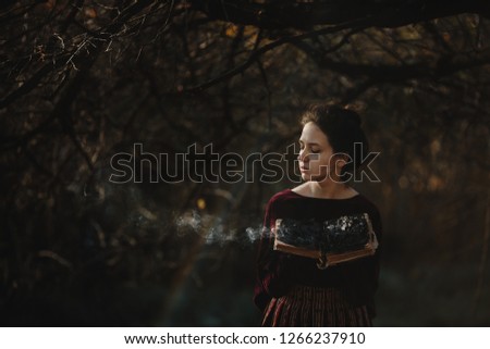 Autumn vibes. 4k. Gothic style. Brunette woman in dark red clothes stands with old burning book under tree with fallen leaves