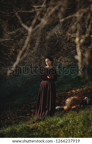 Autumn vibes. 4k. Gothic style. Brunette woman in dark red clothes stands with a pomegranate under tree with fallen leaves