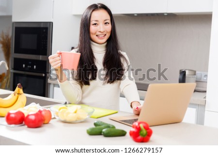 Photo of young asian girl with mug and laptop standing at table with vegetables and fruits
