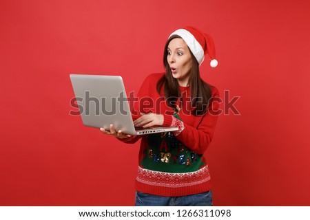 Amazed young Santa girl in Christmas hat looking surprised, working typing on laptop pc computer isolated on red background. Happy New Year 2019 celebration holiday party concept. Mock up copy space