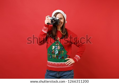 Smiling young Santa girl in sweater, Christmas hat taking pictures on retro vintage photo camera isolated on red background. Happy New Year 2019 celebration holiday party concept. Mock up copy space