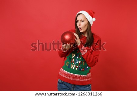 Shocked puzzled young Santa girl looking on big shiny tree toy in hands, keeping mouth wide open isolated on red background. Happy New Year 2019 celebration holiday party concept. Mock up copy space
